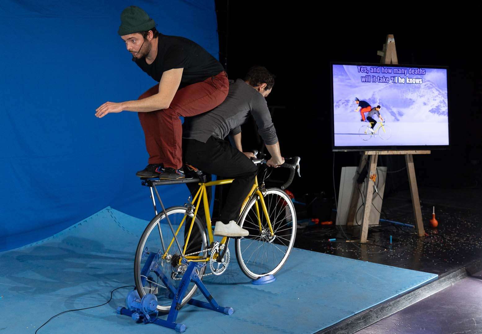 Deux acrobates sont juchés sur un vélo fixe devant un écran bleu